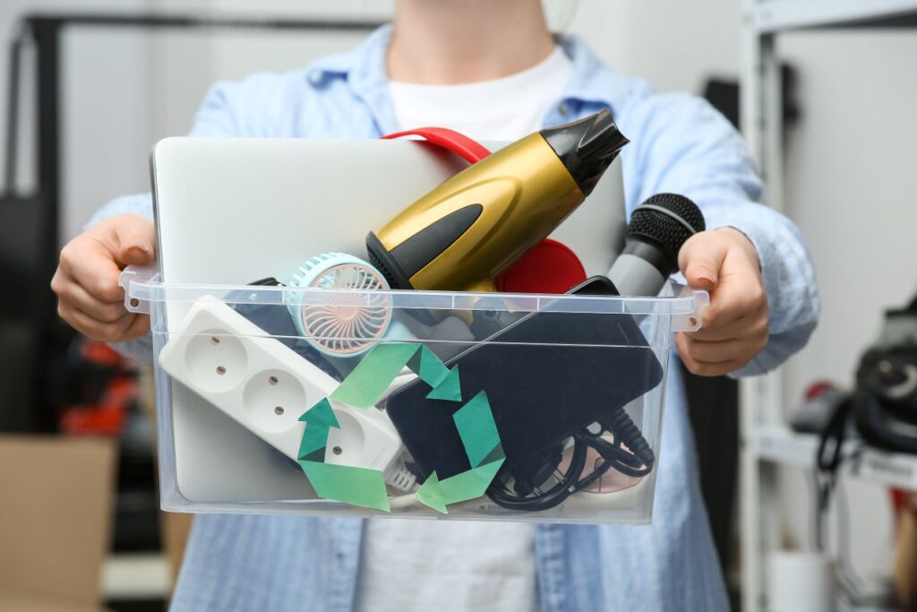 Person haelt eine Kiste mit alten Elektrogeraeten, darunter Foehn, Steckerleiste und Laptop – nachhaltiger Ersatz durch LED Einbaustrahler geplant