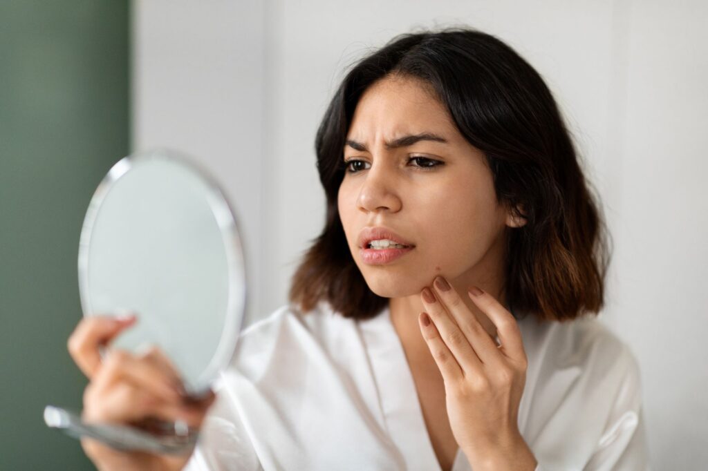  Frau betrachtet ihre Haut im Handspiegel mit skeptischem Blick – Haut und Psyche im Zusammenhang erkennen - Alex Cosmetic
